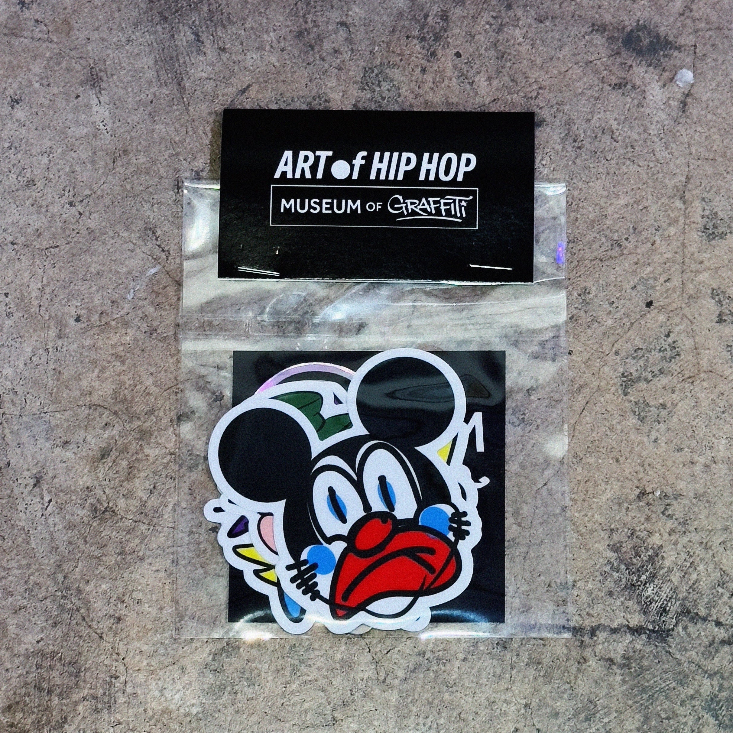 A die-cut sticker featuring a stylized cartoon mouse face with black ears, blue eyes, and a red mouth, shown inside a clear plastic sleeve with a black backing card labeled 'ART of HIP HOP MUSEUM of CHICITY'.